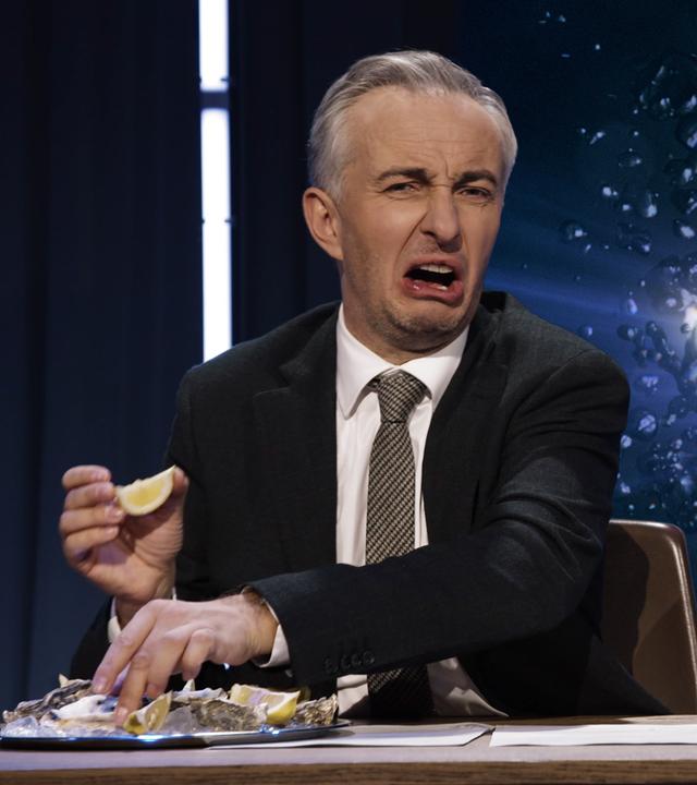 Jan Böhmermann sitzt am Schreibtisch und isst Austern mit einem angeekelten Gesichtsausdruck. Rechts im Hintergrund ist ein leuchtendes Bild einer geöffneten Auster zu sehen.