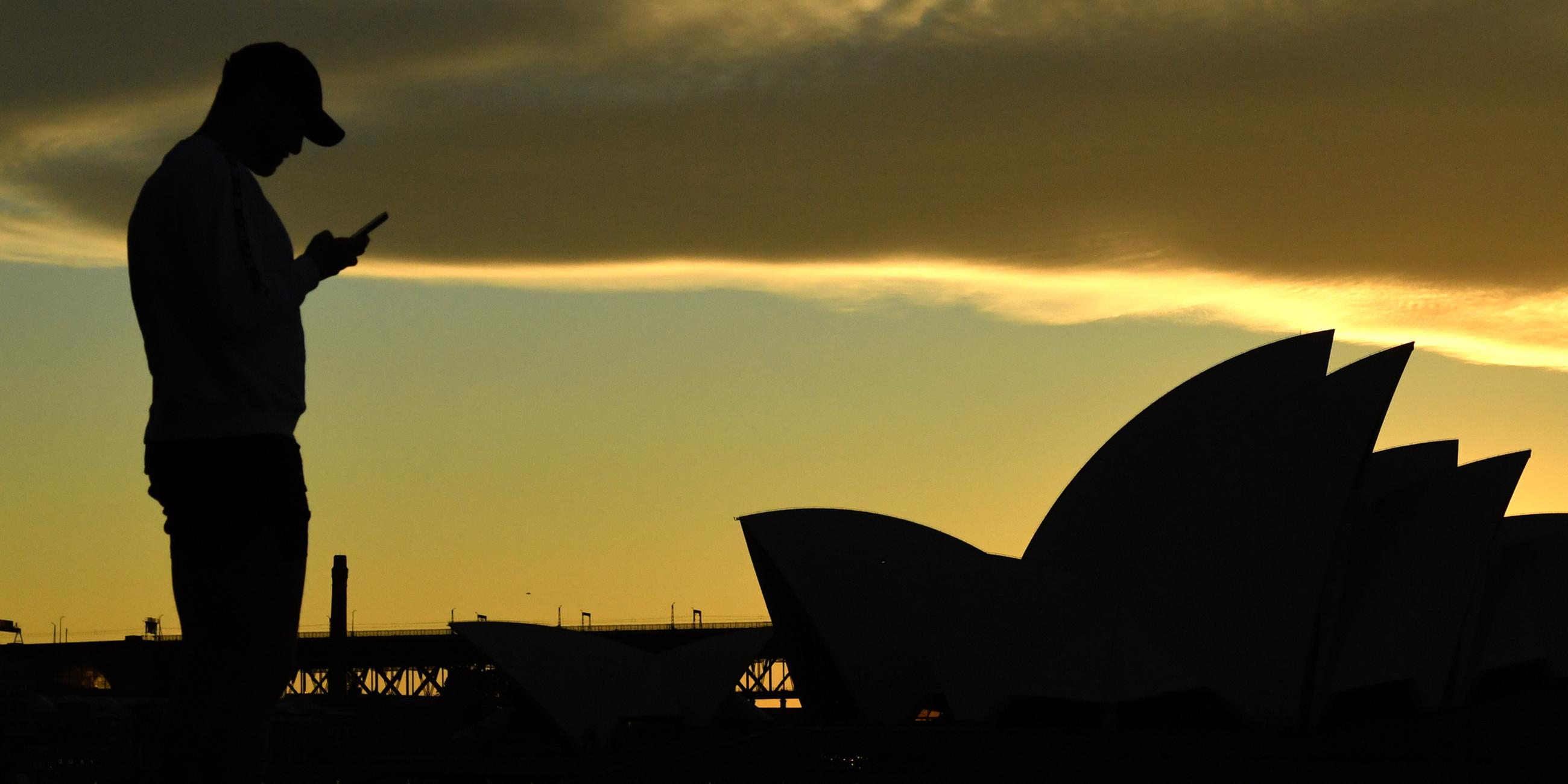 Archiv: Ein Mann steht bei Sonnenuntergang vor dem Sydney Opera House und blickt auf sein Mobiltelefon. (26.07.2021)