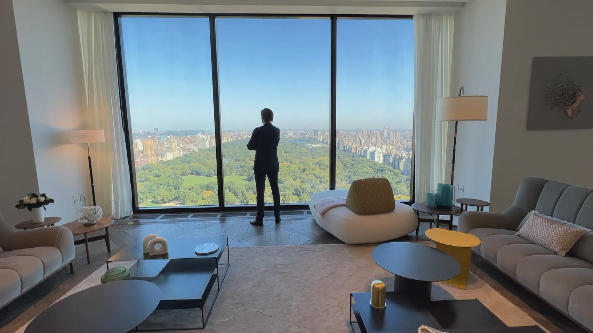 Mann steht in einem luxuriösen Appartemant und schaut durch ein riesiges Panoramafenster auf den Central Park in New York.