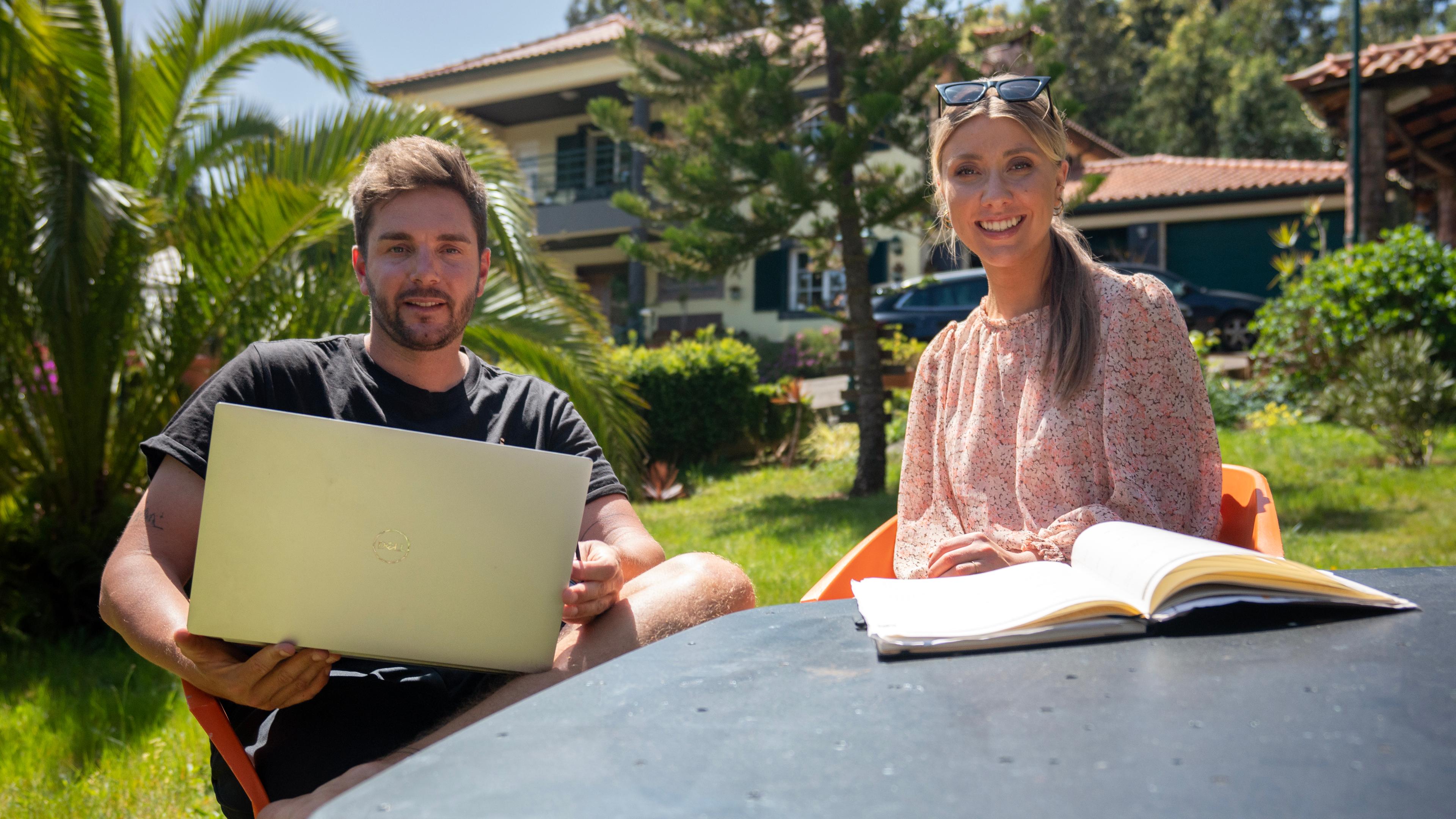Mann und Frau sitzen im Garten am Tisch mit Laptop. Im Hintergrund ist eine Villa mit Palmen.