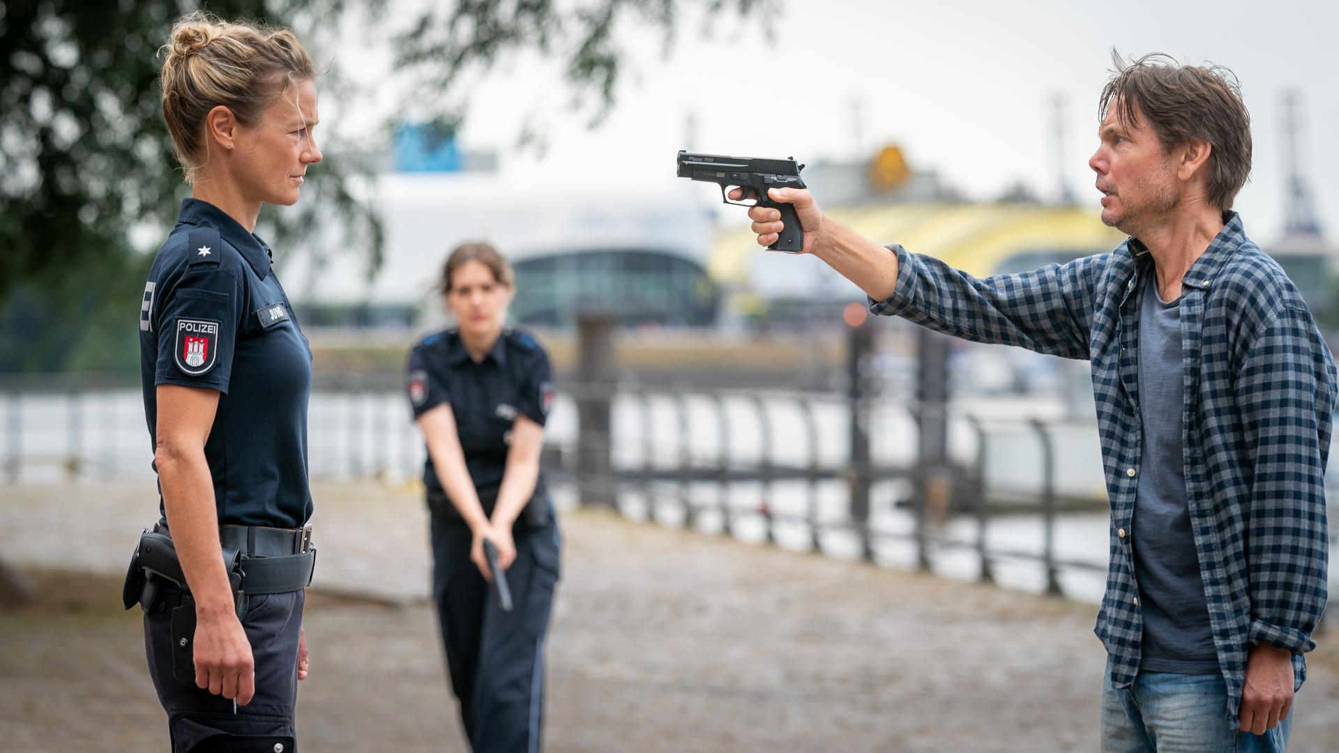 Die Polizistin Franzi Jung (Rhea Harder-Vennewald) und Bernd Oswald (Marek Harloff) stehen sich auf einem Platz gegenüber. Oswald hat seine Waffe auf Franzi gerichtet. Sie ist unbewaffnet. Sie schaut ihn ernst an. Im Hintergrund steht eine Polizistin (Komparsin). Sie hält ihre Waffe schussbereit in den Händen.