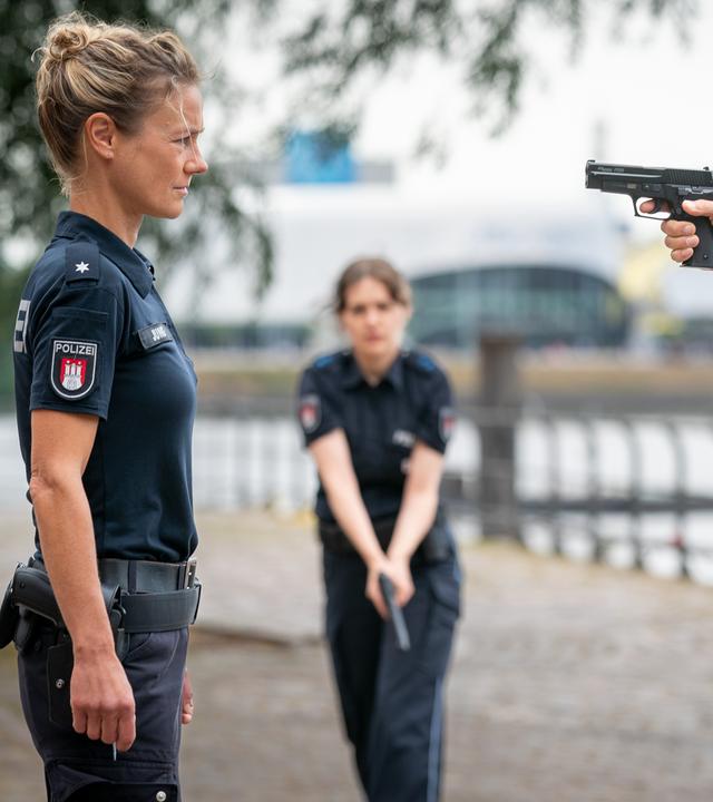 Die Polizistin Franzi Jung (Rhea Harder-Vennewald) und Bernd Oswald (Marek Harloff) stehen sich auf einem Platz gegenüber. Oswald hat seine Waffe auf Franzi gerichtet. Sie ist unbewaffnet. Sie schaut ihn ernst an. Im Hintergrund steht eine Polizistin (Komparsin). Sie hält ihre Waffe schussbereit in den Händen.