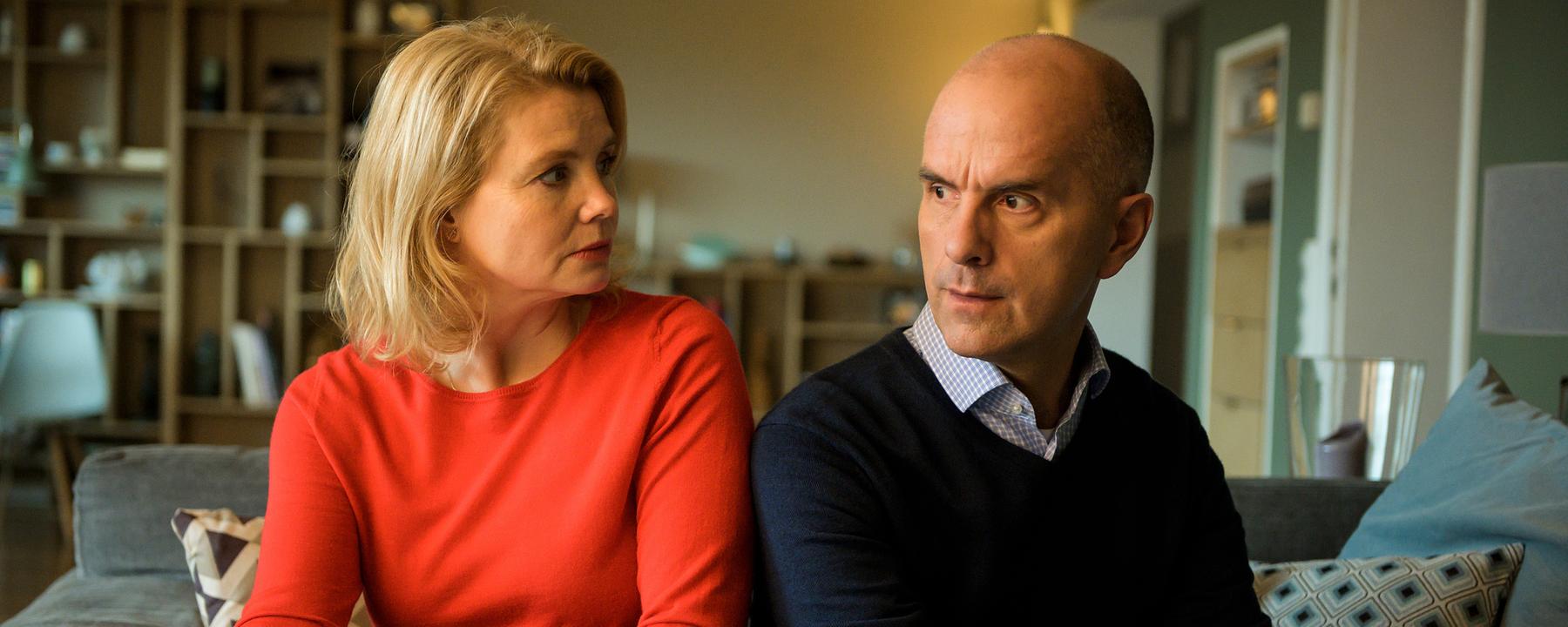 Anne (Annette Frier) sitzt in einem roten Pulli neben ihrem Mann Erik (Christoph Maria Herbst). Sie sitzen nebeneinander in ihrem Wohnzimmer auf der Couch. Beide schauen irritiert.