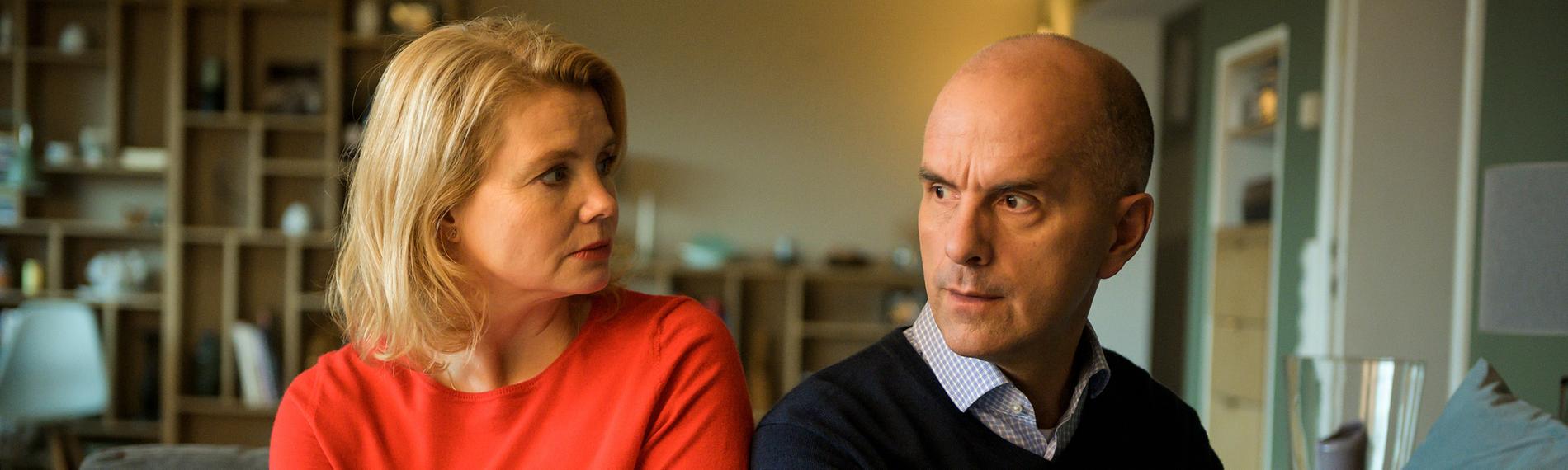 Anne (Annette Frier) sitzt in einem roten Pulli neben ihrem Mann Erik (Christoph Maria Herbst). Sie sitzen nebeneinander in ihrem Wohnzimmer auf der Couch. Beide schauen irritiert.