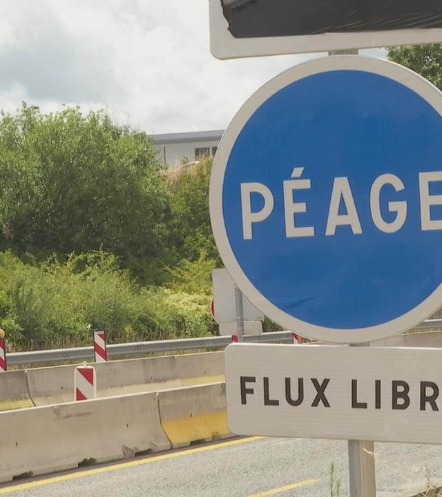 Ein rundes, blaues Straßenschild mit der Aufschrift "PÉAGE" und darunter ein weißes, rechteckiges Schild mit der Aufschrift "FLUX LIBRE". Im Hintergrund sieht man eine Autobahn-Baustelle.
