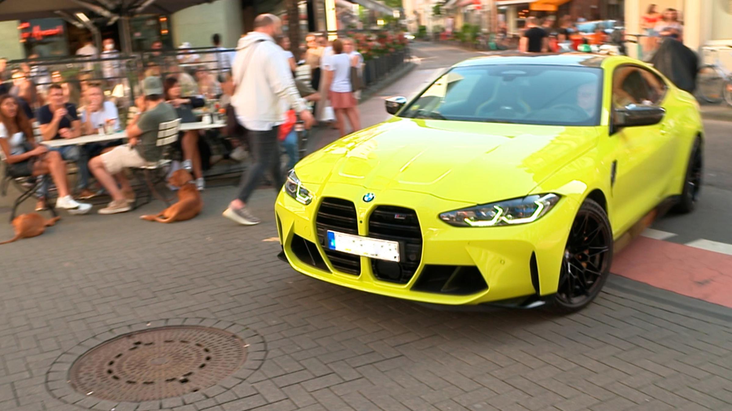 Eine Straßenecke mit voll besetzter Café-Terrasse. Ein knallgelber BMW biegt knapp an den Tischen vorbei um die Ecke.