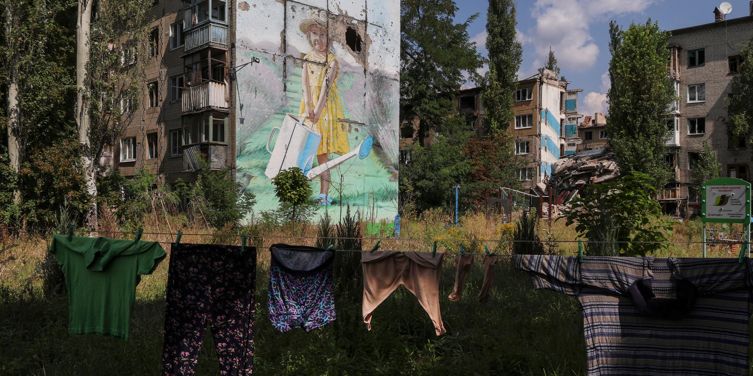 Gewaschene Wäsche trocknet auf einer Leine vor einer beschädigten Wandmalerei in der Siedlung Awdijiwka am 28.08.2023.