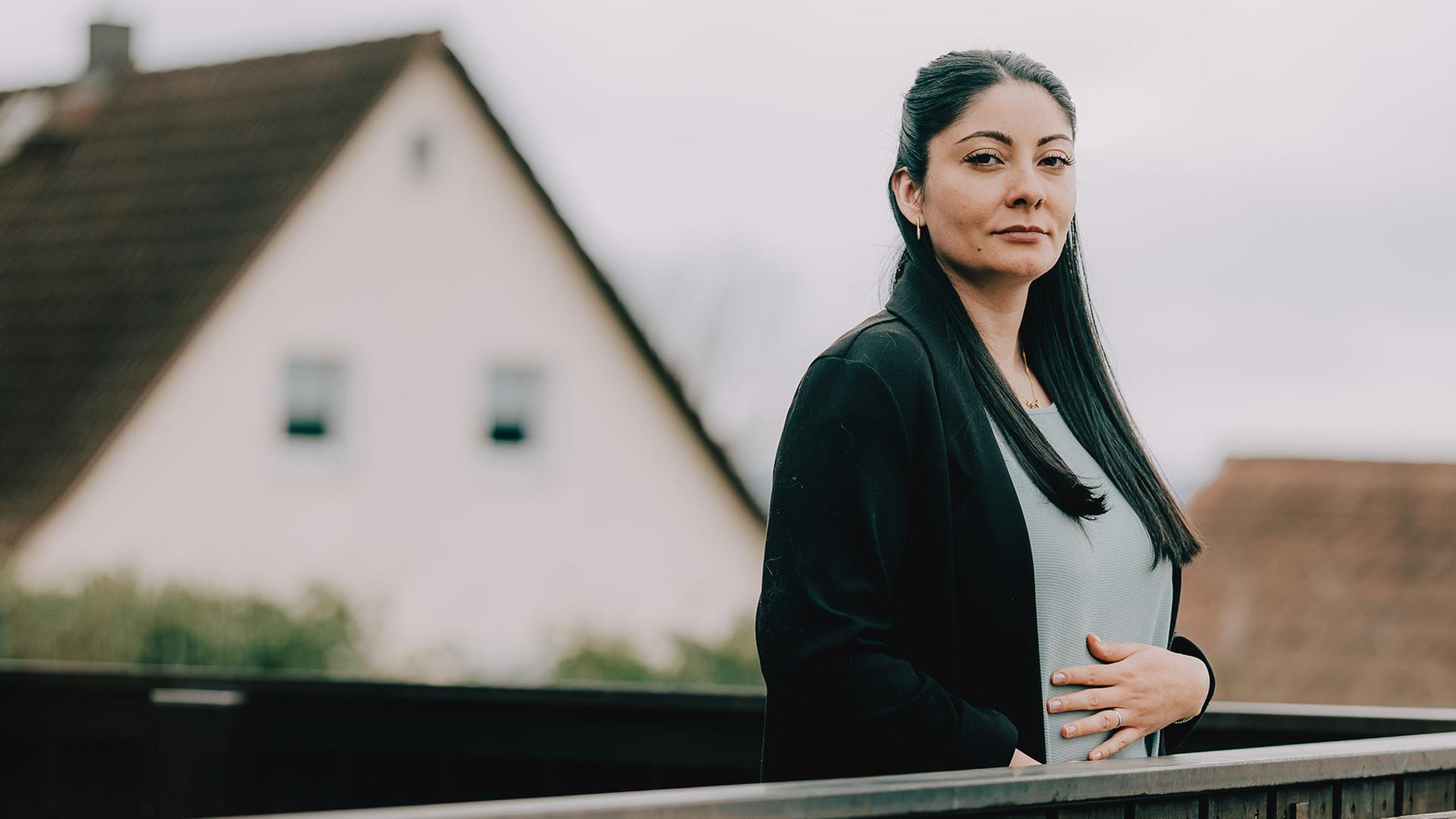 Aylin (rechts im Bild) steht auf einem Balkon. Sie schaut Ernst in die Kamera und hält fasst sich mit der Hand an den Bauch.