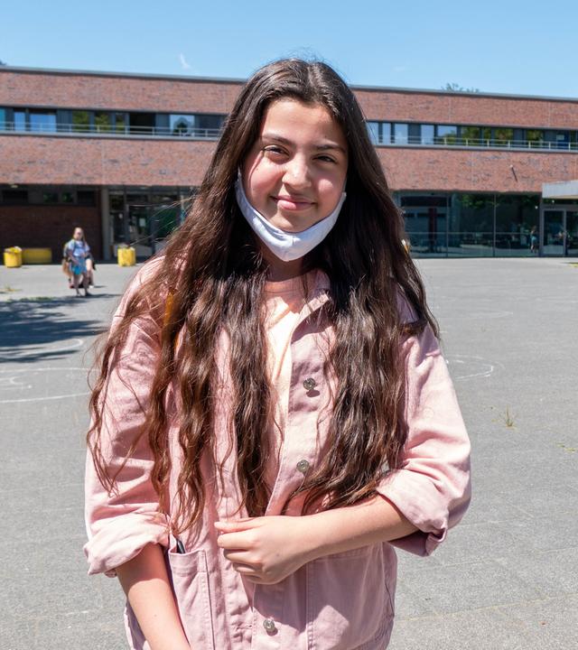 Azra steht mit heruntergezogener Maske vor dem Schulgebäude. Azra hat langes dunkles Haar, trägt eine Jeans und eine rosafarbene Jacke. Der Schulhof hinter ihr ist leer. Sie lacht in die Kamera.