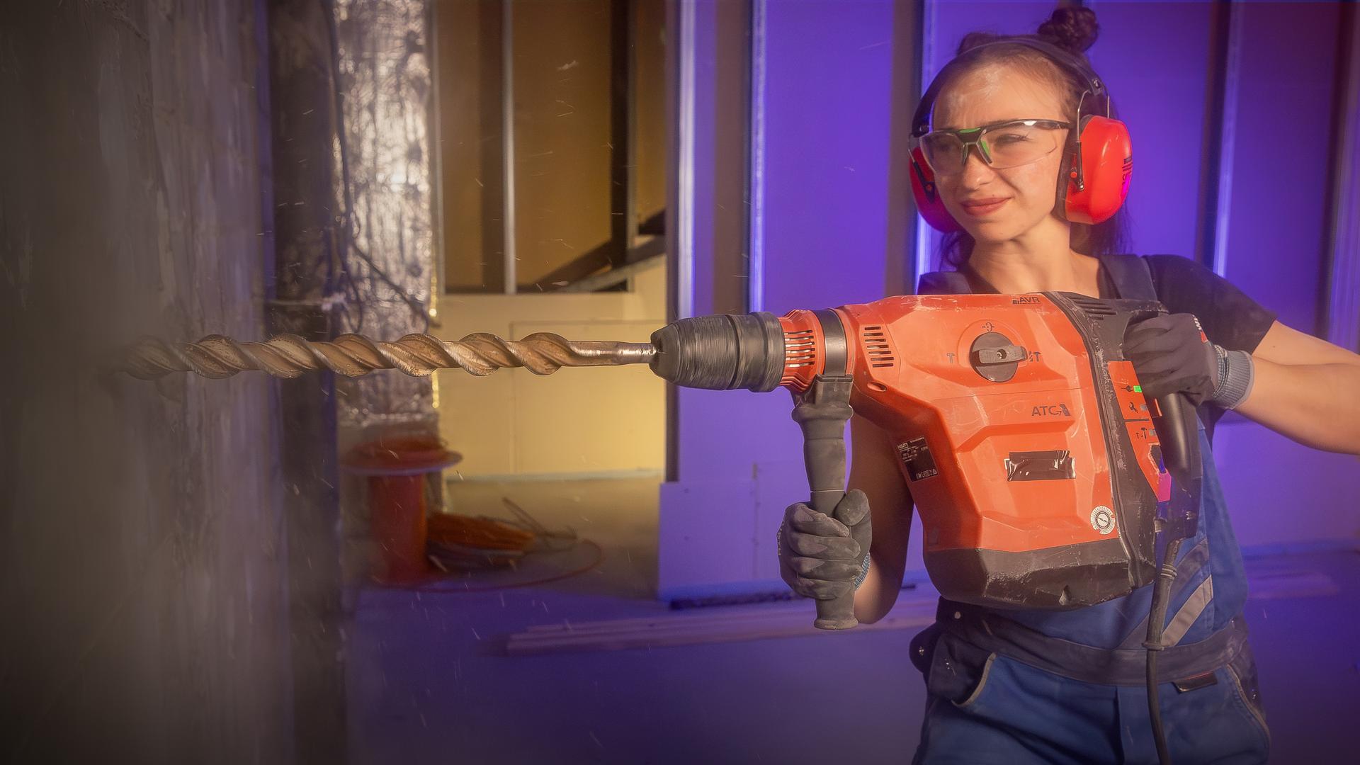 Eine Frau in blauer Arbeitskleidung, Schutzbrille und Gehörschutz bohrt mit einer großen Bohrmaschine in die Wand. Es ist Bohrstaub zu sehen.