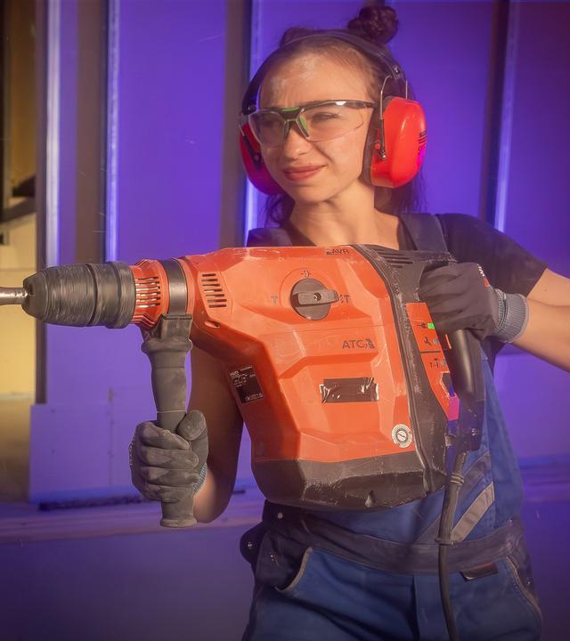 Eine Frau in blauer Arbeitskleidung, Schutzbrille und Gehörschutz bohrt mit einer großen Bohrmaschine in die Wand. Es ist Bohrstaub zu sehen.