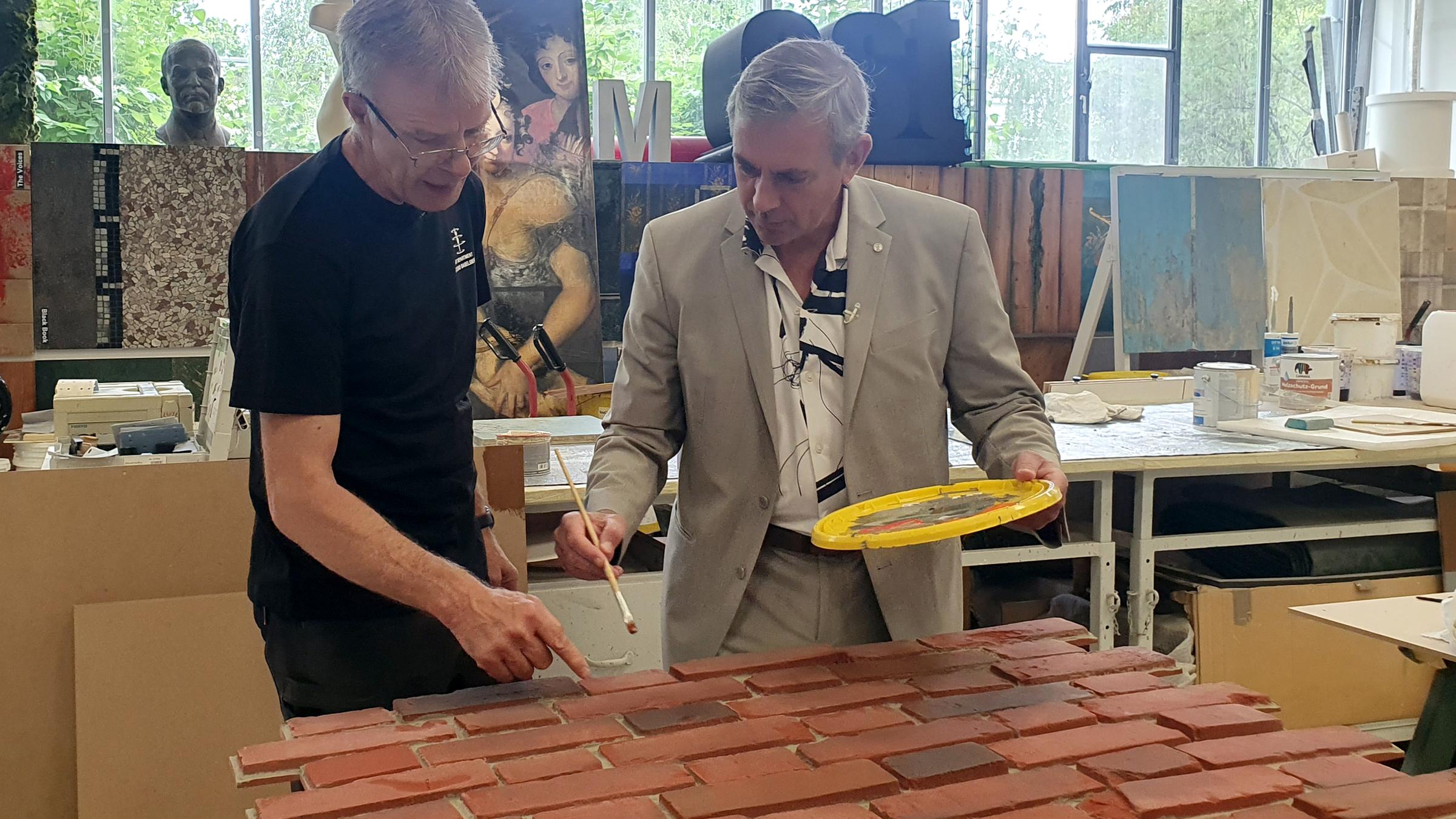 Wladimir Kaminer und Robert Krüger beugen sich über die Attrappe von Klinkersteinen, die sie rot anmalen. Kaminer hält einen Pinsel in der Hand.
