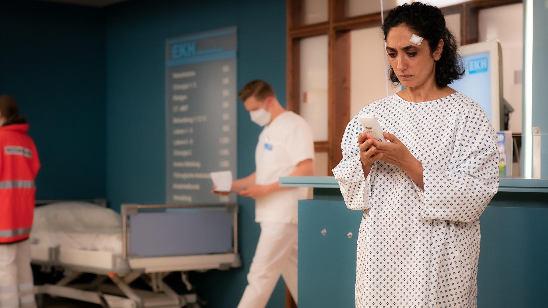 Samira Karimi (Ariella Hirshfeld) steht in einem Patientenhemd vor der Anmeldung im EKH. Sie blickt auf ihr Handy, das sie in den Händen hält.