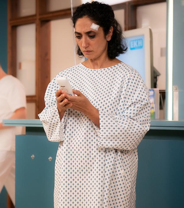 Samira Karimi (Ariella Hirshfeld) steht in einem Patientenhemd vor der Anmeldung im EKH. Sie blickt auf ihr Handy, das sie in den Händen hält.