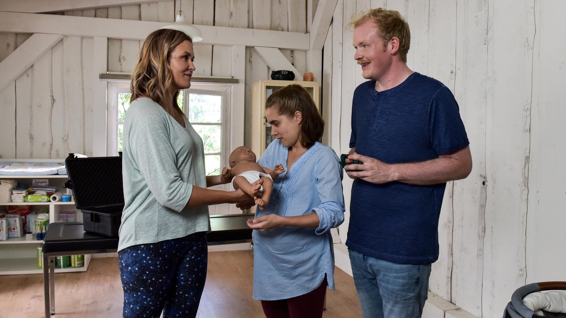 "Lena Lorenz - Baby auf Probe": In der Hebammenpraxis überreicht Lena Lorenz (Judith Hoersch) der schwangeren Alina Moosleitner (Anne-Marie Lux) eine Babypuppe. Daneben steht Tim Scheuer (Sebastian Griegel) und schaut freudig.