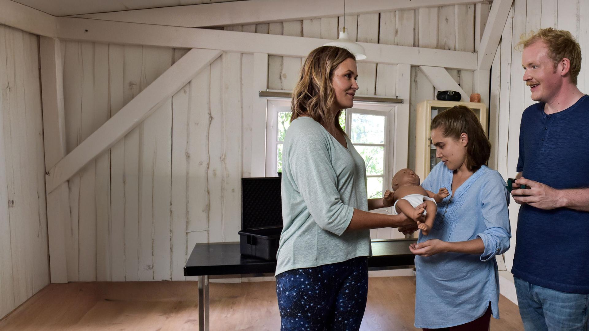 "Lena Lorenz - Baby auf Probe": In der Hebammenpraxis überreicht Lena Lorenz (Judith Hoersch) der schwangeren Alina Moosleitner (Anne-Marie Lux) eine Babypuppe. Daneben steht Tim Scheuer (Sebastian Griegel) und schaut freudig.