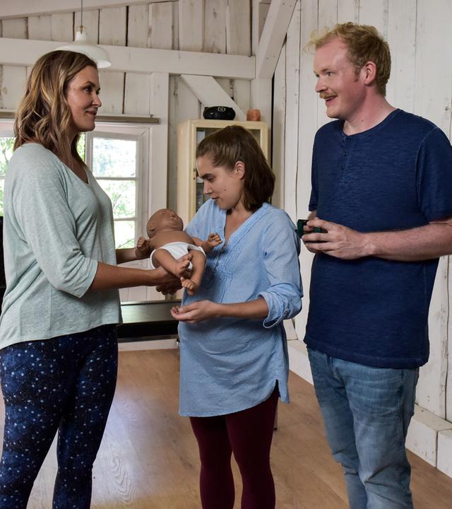 "Lena Lorenz - Baby auf Probe": In der Hebammenpraxis überreicht Lena Lorenz (Judith Hoersch) der schwangeren Alina Moosleitner (Anne-Marie Lux) eine Babypuppe. Daneben steht Tim Scheuer (Sebastian Griegel) und schaut freudig.