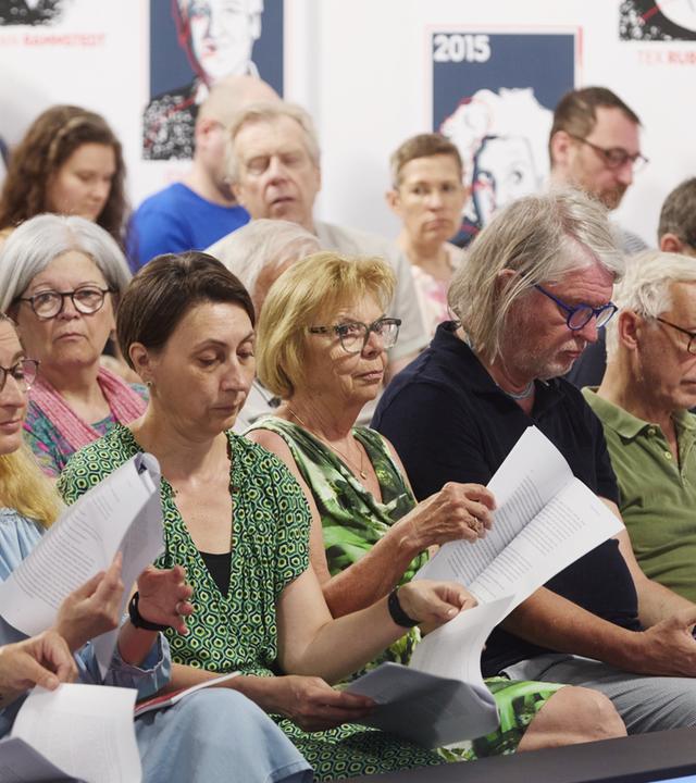Das Bild zeigt eine Veranstaltung im Rahmen der 50. Tage der deutschsprachigen Literatur, speziell den Ingeborg-Bachmann-Preis 2026. Im Vordergrund sitzen mehrere Personen in Reihen auf Stühlen. Die Zuhörer halten Papiere in den Händen.