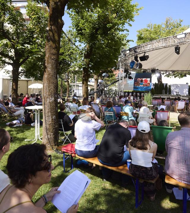 Das Bild zeigt eine Veranstaltung während der 50. Tage der deutschsprachigen Literatur, dem Ingeborg-Bachmann-Preis 2026. Es ist ein sonniger Tag, und die Szene findet im Freien statt, umgeben von Bäumen.