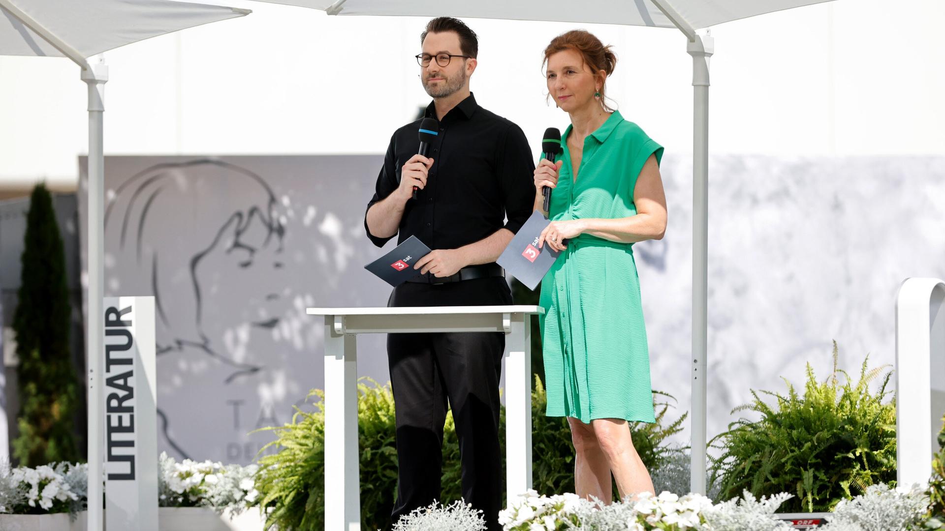Zwei Moderator:innen stehen unter weißen Sonnenschirmen auf einer Bühne im Freien und sprechen in Mikrofone; im Hintergrund ist das Logo der Tage der deutschsprachigen Literatur sichtbar.