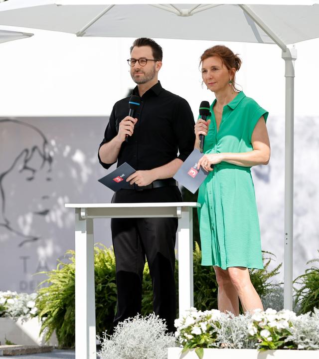 Zwei Moderator:innen stehen unter weißen Sonnenschirmen auf einer Bühne im Freien und sprechen in Mikrofone; im Hintergrund ist das Logo der Tage der deutschsprachigen Literatur sichtbar.