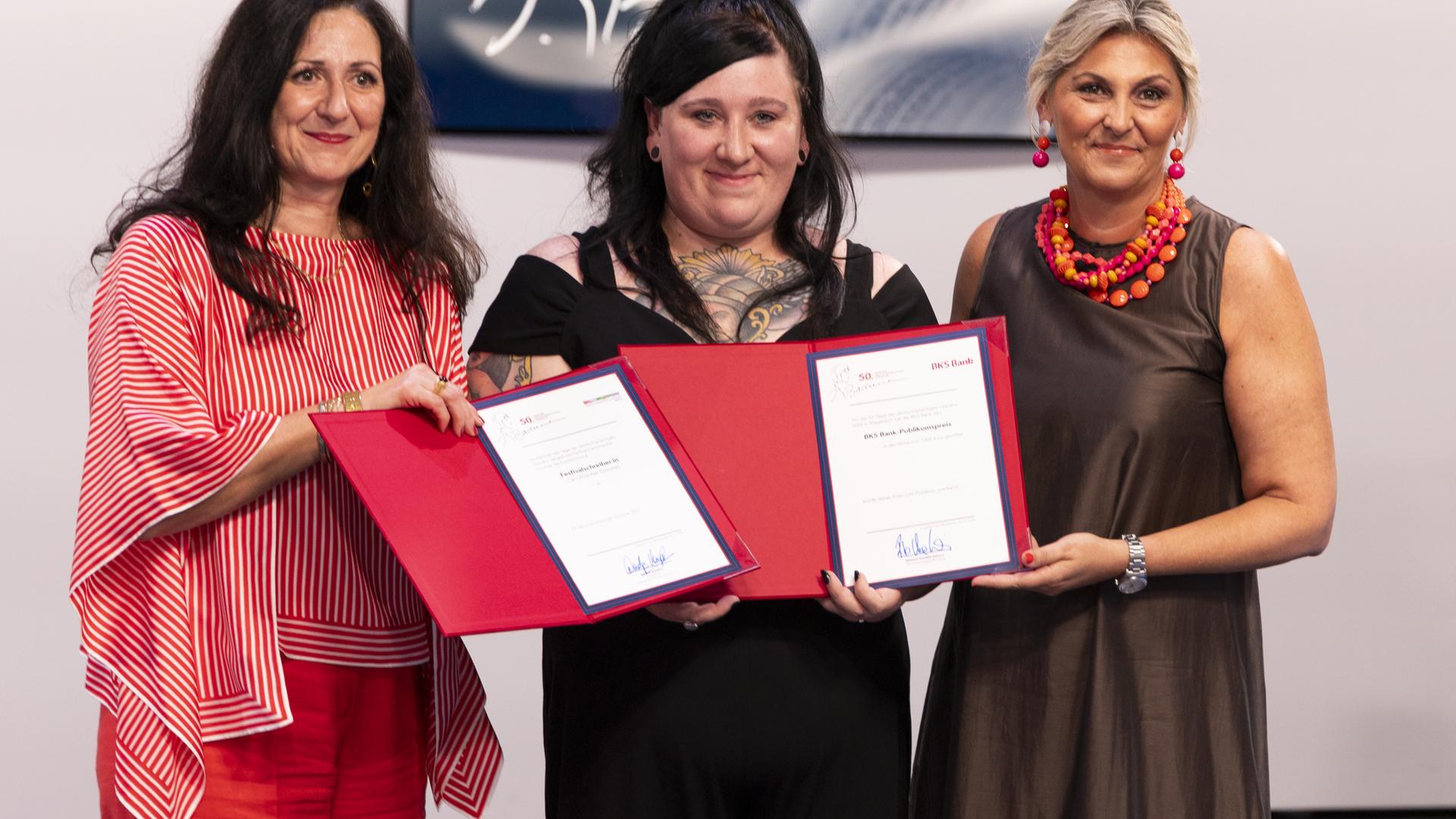 Drei Frauen stehen nebeneinander und präsentieren glücklich lächelnd zwei rote Kladden mit zwei Preisurkunden.