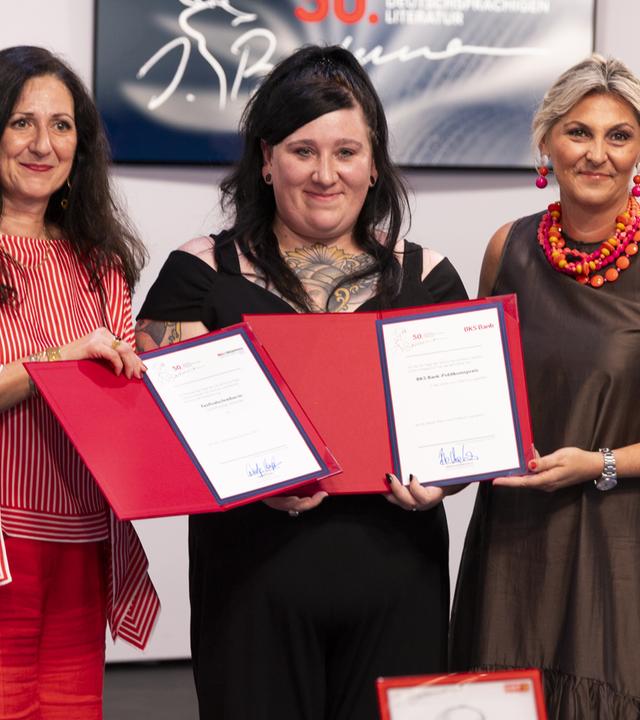Drei Frauen stehen nebeneinander und präsentieren glücklich lächelnd zwei rote Kladden mit zwei Preisurkunden.
