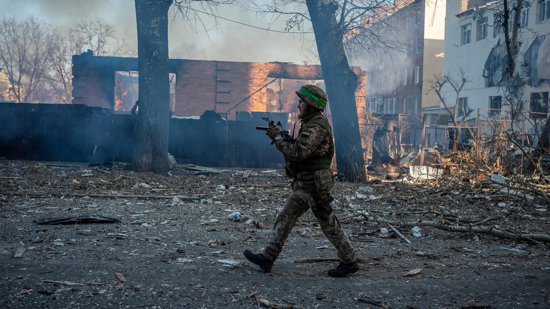 Ein ukrainischer Soldat läuft über zerstörte Straße. Hinter ihm zertrümmerte, verbrannte Häuser. 
