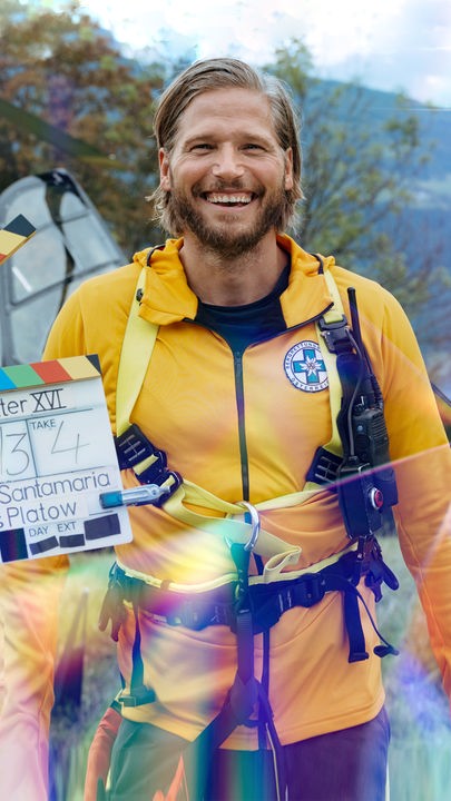 Sebastian Ströbel aus der Serie "Die Bergretter" steht vor einem Rettungs-Helikopter. Vor ihm sieht man eine Filmklappe, denn das Foto entstand während der Dreharbeiten. Die Doku Backstage schaut hinter die Kulissen der Serie.
