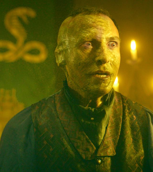 Ein im Gesicht geschuppter Mann schaut mit großen Augen nach rechts. Links von ihm im Hintergrund ist ein Schlangensymbol zu sehen.