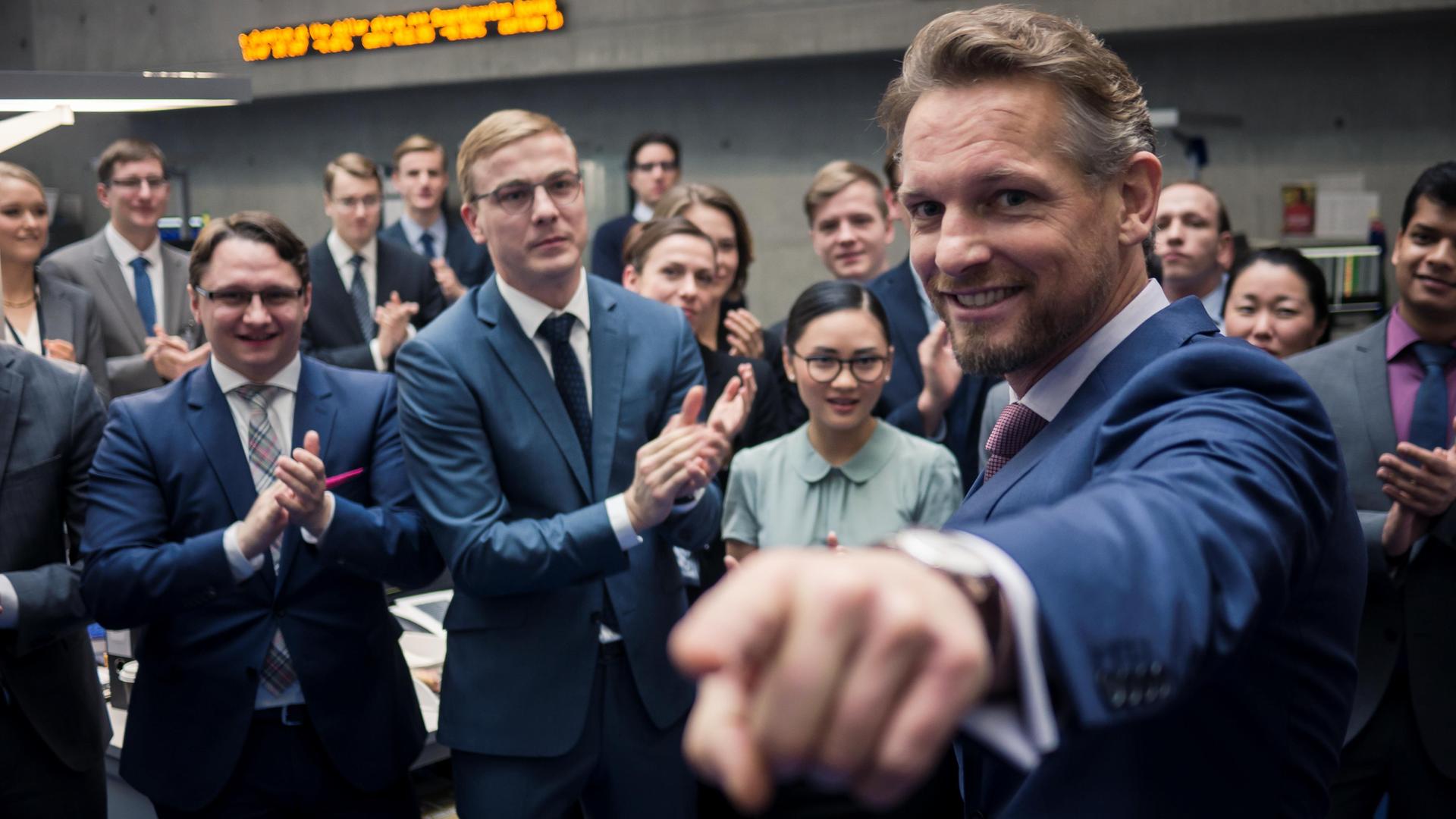 Eine Gruppe voller Menschen klatscht Barry Atsma (Gabriel Fenger) Beifall. Dieser lächelt in die Kamera und deutet mit dem Zeigefinger in Kamerarichtung.