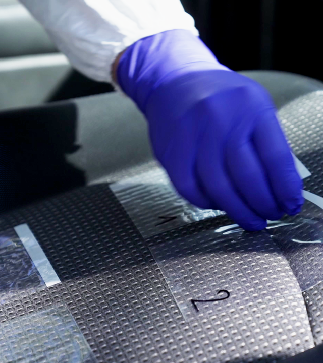 Hand mit Einmalhandschuh bekleidet klebt Klebestreifen auf einen Autositz. Spurensicherung. Der Klebestreifen trägt die mit schwarzem Edding aufgetragene Ziffer 2. Links daneben: Lichtschematische Darstellung eines Fingerabdrucks.