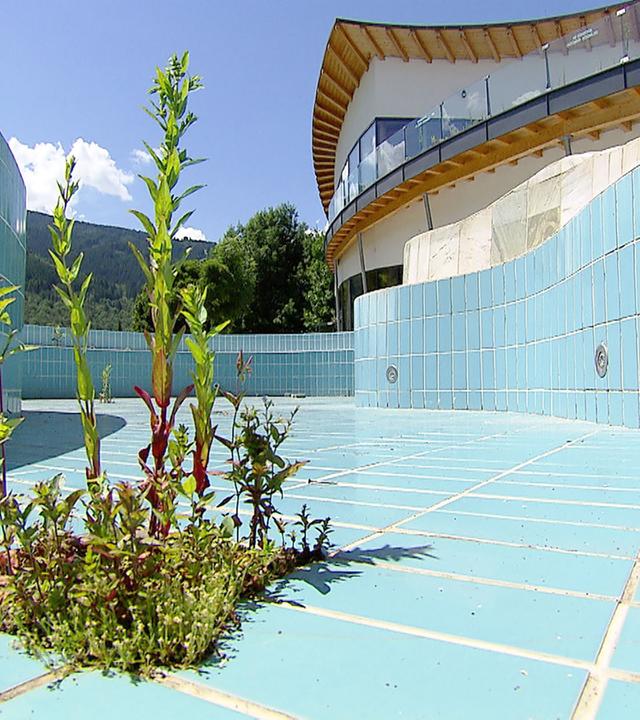 Das Bild zeigt ein leer stehendes Schwimmbecken in einem Hallenbad. Die Fliesen des Beckens sind in einem hellen Blau gehalten und zeigen einige Abnutzungserscheinungen. In der Mitte des Beckens sind grüne Pflanzen zu sehen, die aus den Fugen der Fliesen wachsen, was auf Verwahrlosung hinweist. Die Wände des Schwimmbeckens sind rundlich und dekoriert, während sich hinter dem Becken ein modernes Gebäude mit einer großen Fensterfront und einem geschwungenen Dach befindet. Der Himmel ist blau mit einigen weißen Wolken, und im Hintergrund sind grüne Berge sichtbar. Es gibt keine Menschen im Bild, was die verlassene Atmosphäre des Ortes verstärkt.