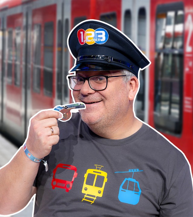 Elton steht mit Trillerpfeife in der Hand und einer Lokführermütze vor einem Zug. 