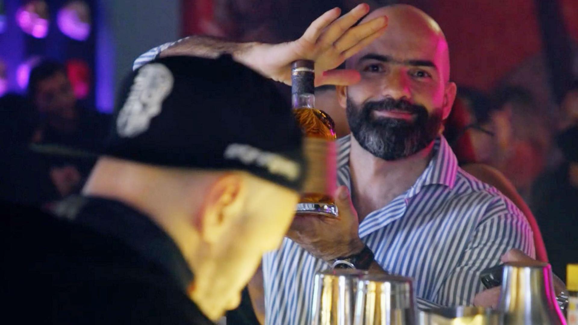 Drei Männer an einer Bar. Der bärtige Mann in der Mitte blickt direkt in die Kamera und präsentiert mit beiden Händen eine Flasche mit einem alkoholischen Getränk.