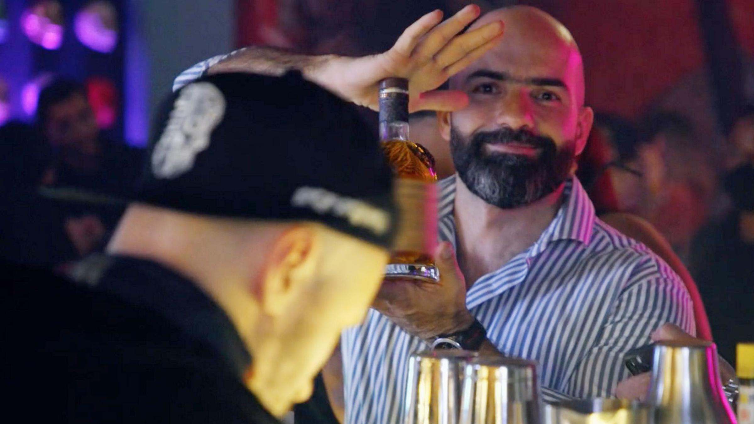 Drei Männer an einer Bar. Der bärtige Mann in der Mitte blickt direkt in die Kamera und präsentiert mit beiden Händen eine Flasche mit einem alkoholischen Getränk.