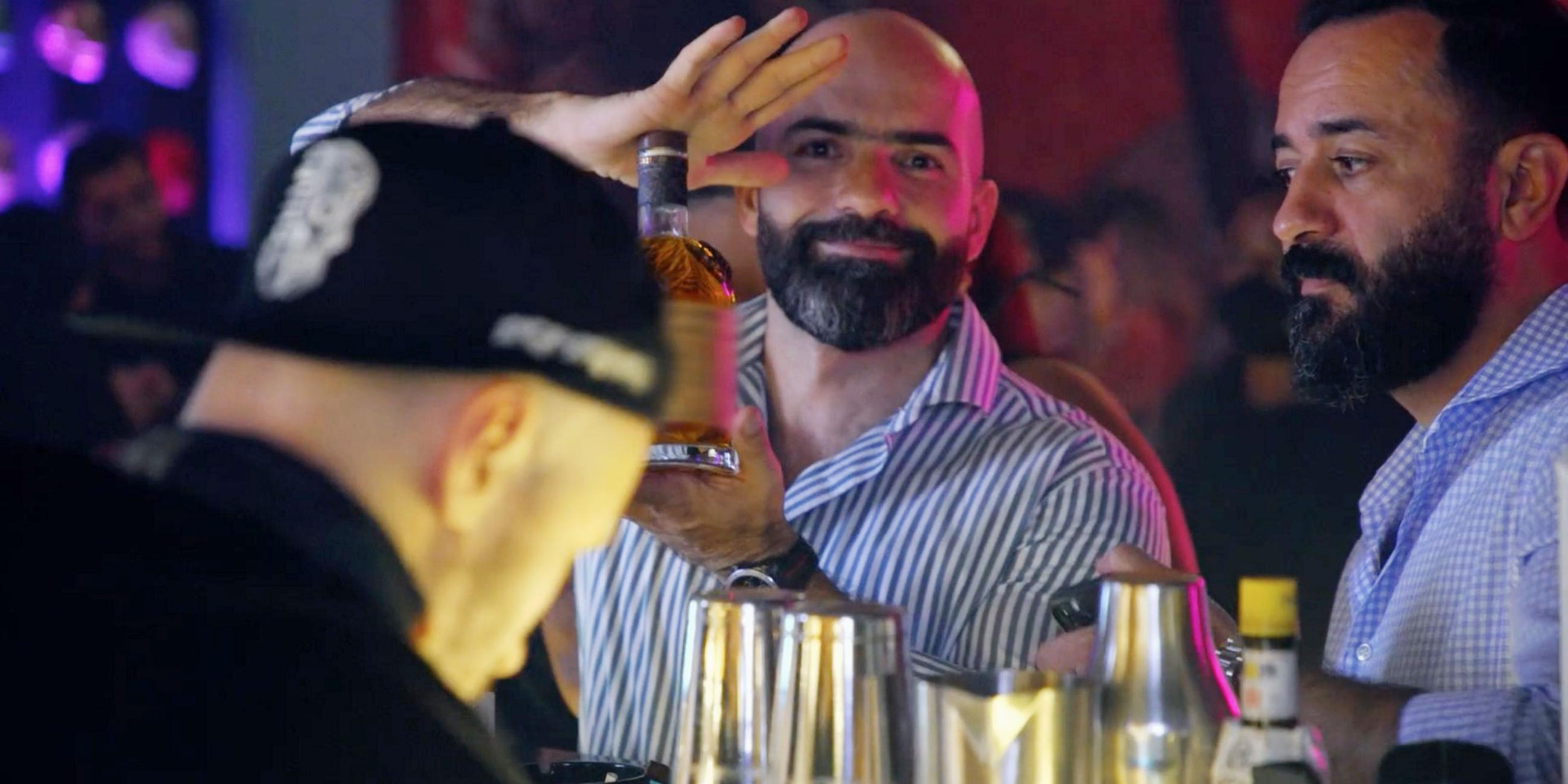 Drei Männer an einer Bar. Der bärtige Mann in der Mitte blickt direkt in die Kamera und präsentiert mit beiden Händen eine Flasche mit einem alkoholischen Getränk.