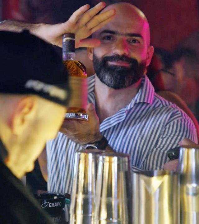 Drei Männer an einer Bar. Der bärtige Mann in der Mitte blickt direkt in die Kamera und präsentiert mit beiden Händen eine Flasche mit einem alkoholischen Getränk.