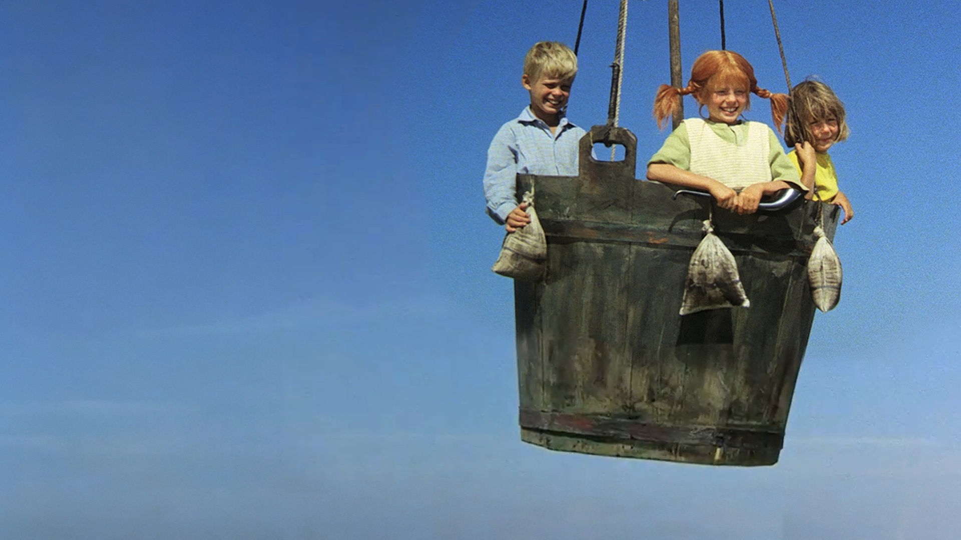 Tommy, Pippi und Annika stehen in einem großen Korb. Sie schweben mit dem Ballon in der Luft und schauen nach unten.