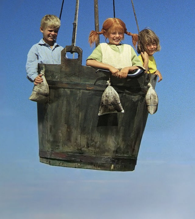 Tommy, Pippi und Annika stehen in einem großen Korb. Sie schweben mit dem Ballon in der Luft und schauen nach unten.
