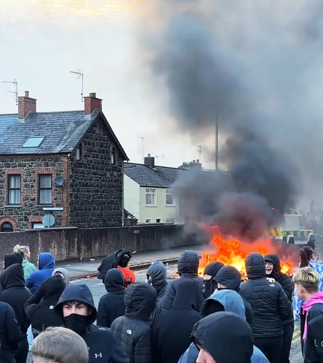 Eine große Gruppe vermummter Menschen steht auf einer Straße, auf der etwas brennt. Hinter dem Feuer sind Einsatzfahrzeuge zu sehen.