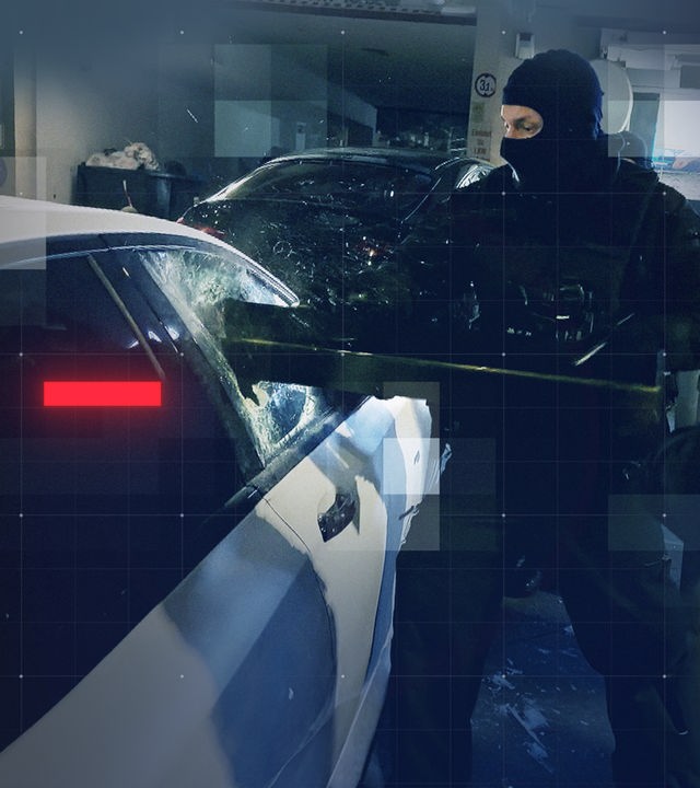 Zwei Vermummte Polizisten stehen neben einem geparktem Pkw, in dem sich mutmaßlich eine Waffe befindet. Einer von ihnen öffnet die Seitenscheibe des Fahrzeuges gewaltsam mit einer Ramme
