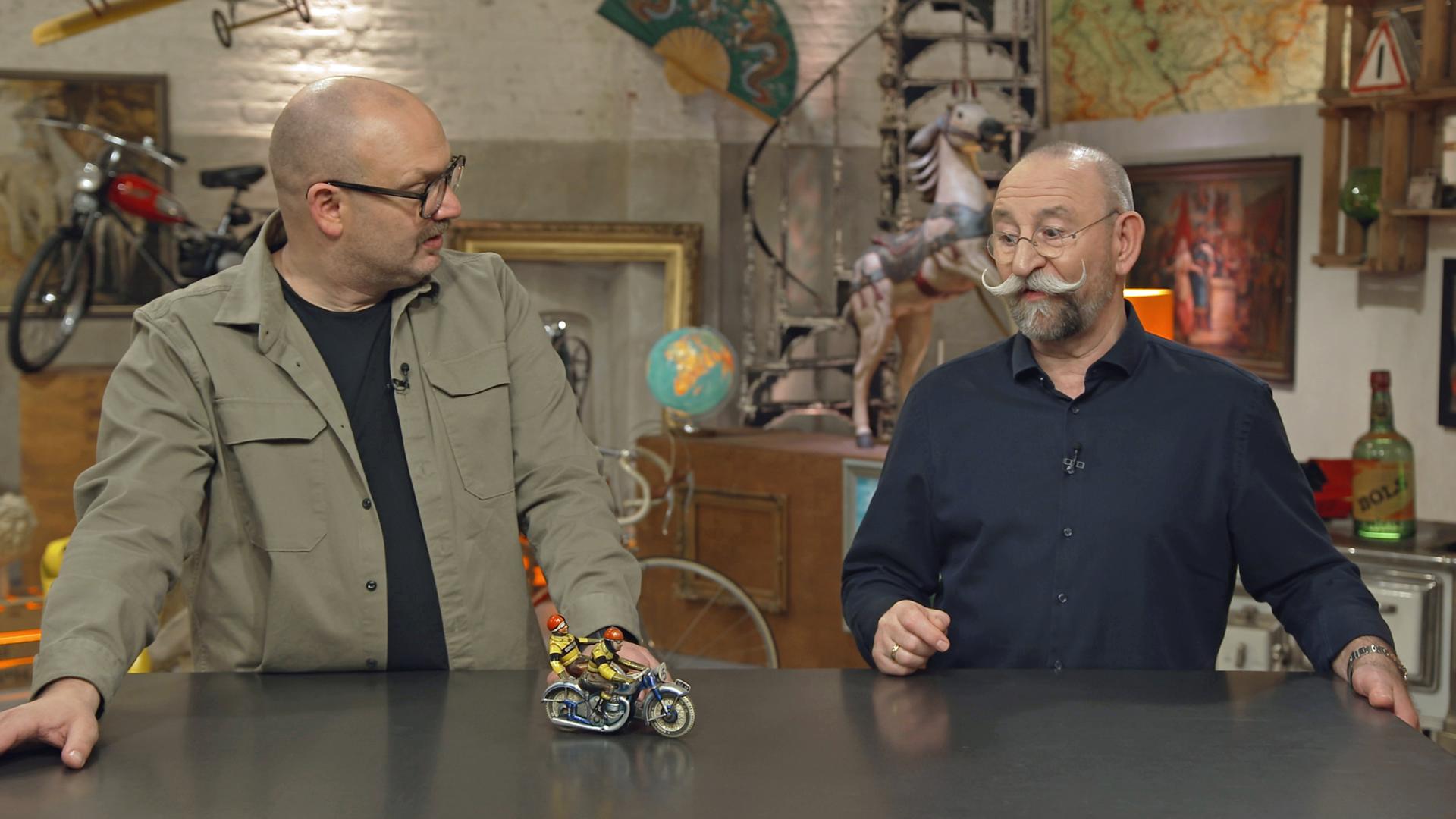 Sven Deutschmanek und Horst Lichter stehen am Expertisentisch, worauf ein kleines Motorrad-Blechspielzeug zu sehen ist.
