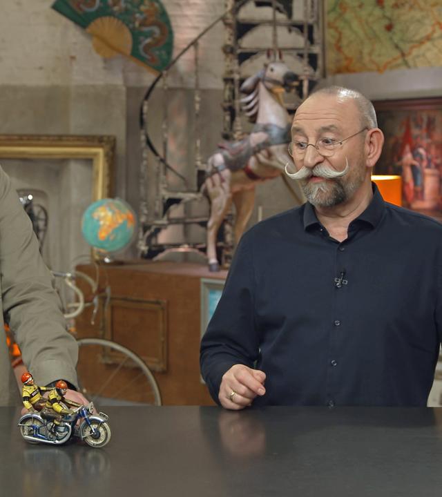 Sven Deutschmanek und Horst Lichter stehen am Expertisentisch, worauf ein kleines Motorrad-Blechspielzeug zu sehen ist.