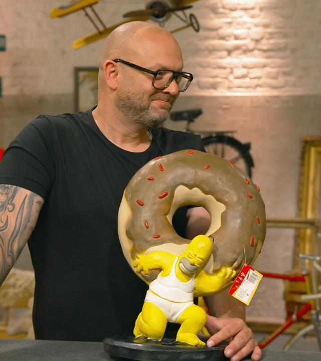Sven Deutschmanek steht grinsend hinter einer Latex-Figur von Homer Simpson. Dieser stemmt einen übergroßen Donut.