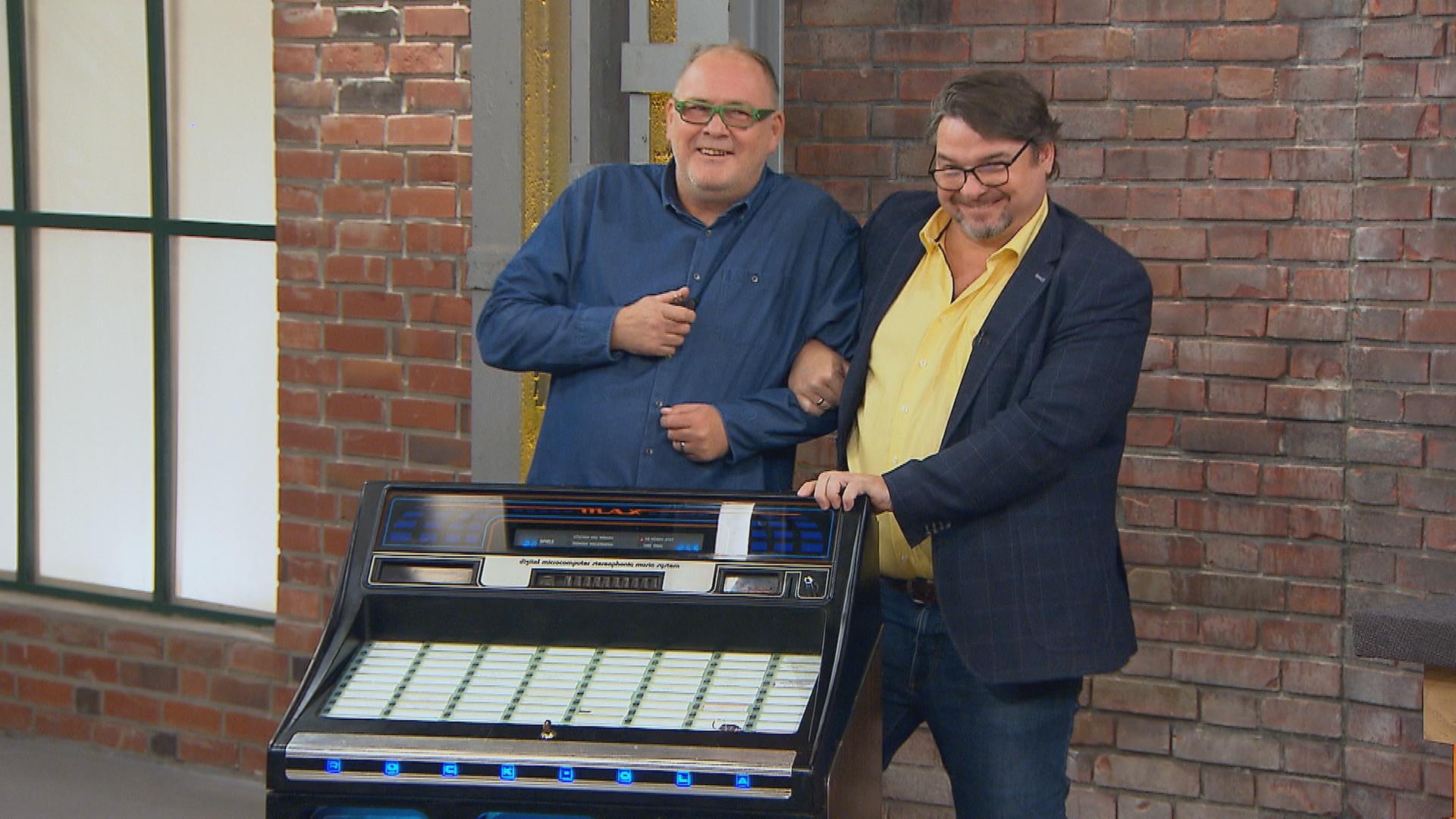 Die Händler Walter "Waldi" Lehnertz und Daniel Meyer stehen zusammen hinter einer Musikbox und tanzen.