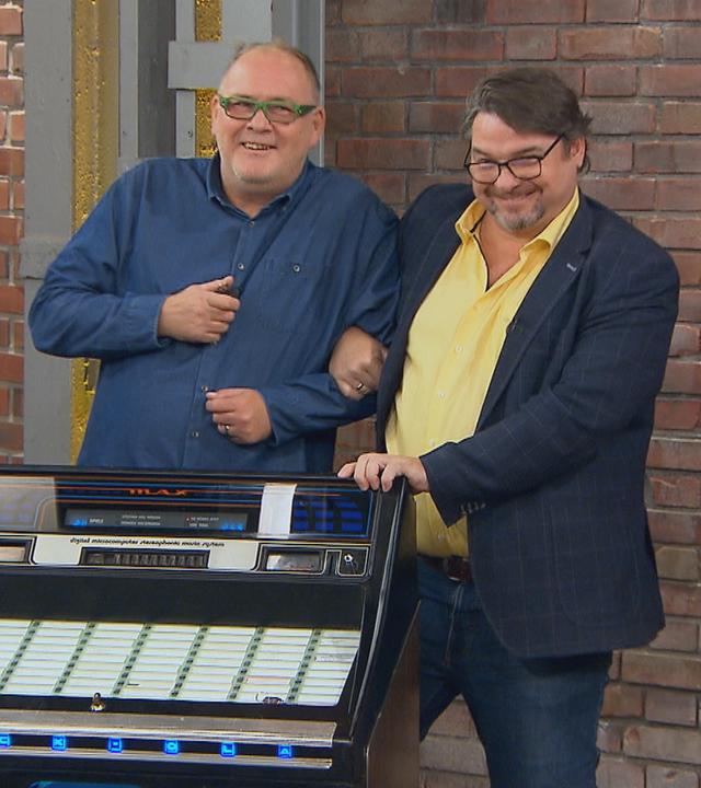 Die Händler Walter "Waldi" Lehnertz und Daniel Meyer stehen zusammen hinter einer Musikbox und tanzen.