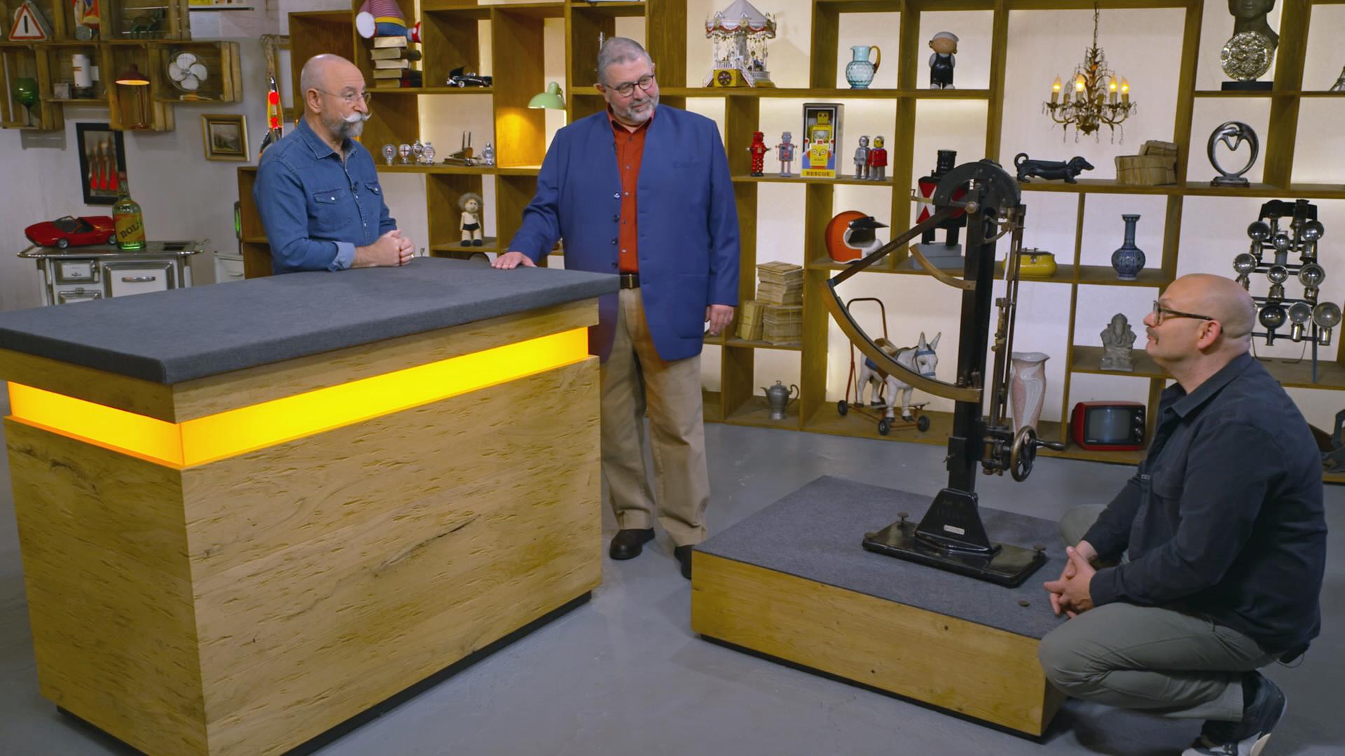 Horst Lichter steht neben einem älteren Verkäufer am Expertisentisch. Sven Deutschmanek kniet neben einem Fertigkeitsprüfgerät und schaut es sich genauer an.