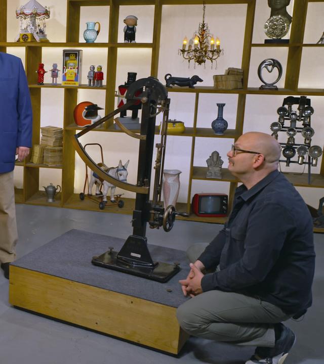 Horst Lichter steht neben einem älteren Verkäufer am Expertisentisch. Sven Deutschmanek kniet neben einem Fertigkeitsprüfgerät und schaut es sich genauer an.