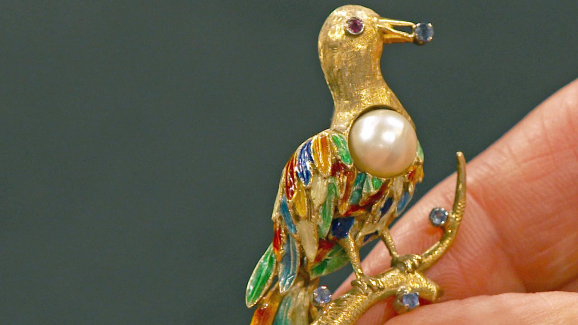 Ein goldene Brosche in Form eines Paradiesvogels mit einer Perle und kleinen, bunten Edelsteinen